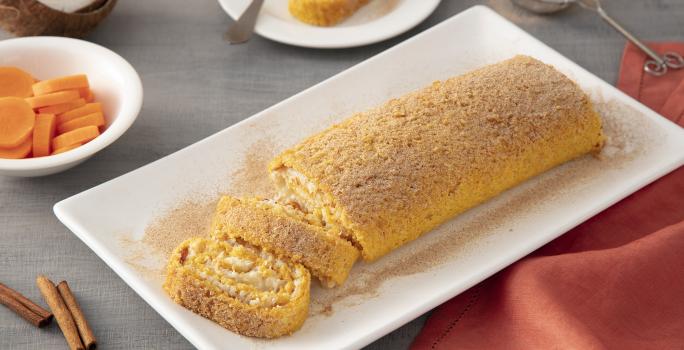 Foto da Receita de Rocambole de Cenoura. Observa-se o rocambole em um prato quadrado branco em fatias. Atrás tem um prato de sobremesa com uma fatia, cenouras em rodelas e canela em pau decoram a foto