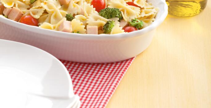 Fotografia em tons de amarelo em uma mesa de madeira com um pano quadriculado em vermelho, um recipiente oval branco com a salada de macarrão. Ao lado, um potinho com azeite.