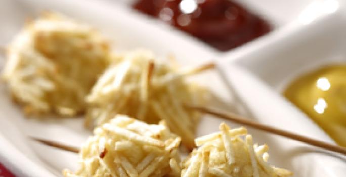 Fotografia em tons de vermelho em uma mesa com uma toalha vermelha com bolinhas brancas, um recipiente branco com o espetinho de cachorro quente empanado com batata palha, e ketchup e mostarda ao lado.