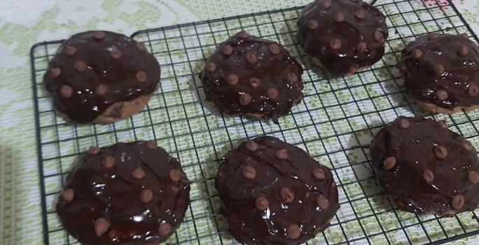 Foto da receita de Cookies low carb de chocolate. Observa-se uma grade preta com 7 cookies dispostos em cima, com calda de chocolate e chocolate em gotas na cobertura.