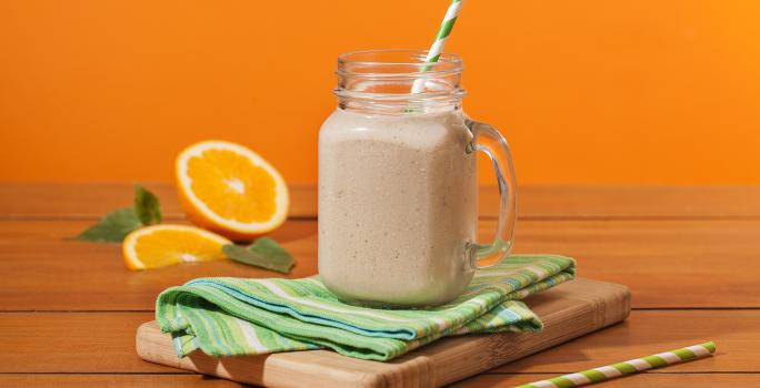 Fotografia em tons de laranja em uma bancada de madeira, um fundo laranja. Ao centro, uma tábua de madeira, um paninho verde, um canudo de papel verde e em cima um copo grande de vidro com o shake de banana dentro. Ao fundo, uma laranja aberta e folhas.