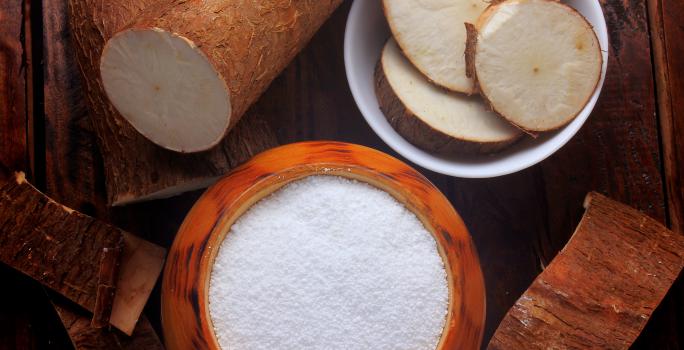 fotografia em tons de marrom e branco de uma bancada marrom vista de cima, contém um recipiente redondo marrom com a farinha dentro e ao lado um recipiente redondo branco com pedaços de mandioca. Em volta contém mandiocas.