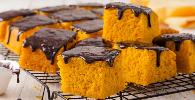 Fotografia em tons de amarelo e preto de uma bancada de madeira com um potinho branco com calda de chocolate, ao lado uma grade preta com fatias quadradas de bolo de cenoura com calda de chocolate em cima, ao fundo, um pano amarelo.