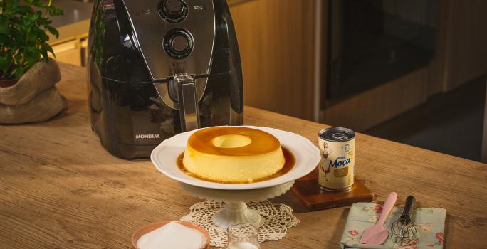 Fotografia em tons de amarelo em uma bancada de madeira, um suporte para bolo branco e o pudim e Leite Moça em cima dele. Ao fundo, uma air fryer Mondial, uma lata de leite Moça, um paninho azul florido com uma espátula rosa e um fouet em cima.