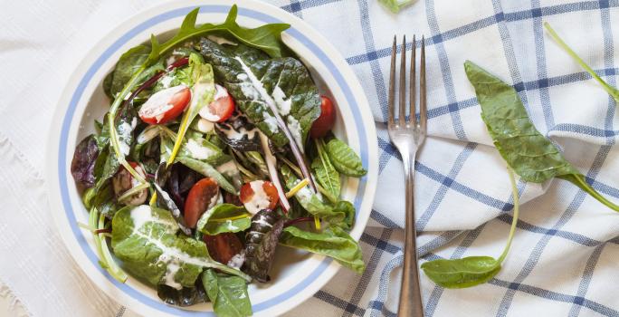 Fotografia vista de cima de um recipiente fundo branco com uma listra azul claro na borda, com uma salada de folhas, com molho de creme de leite e tomates cereja. O recipiente está sobre duas toalhas de mesa, nas cores branco e azul, e ao lado, um garfo.