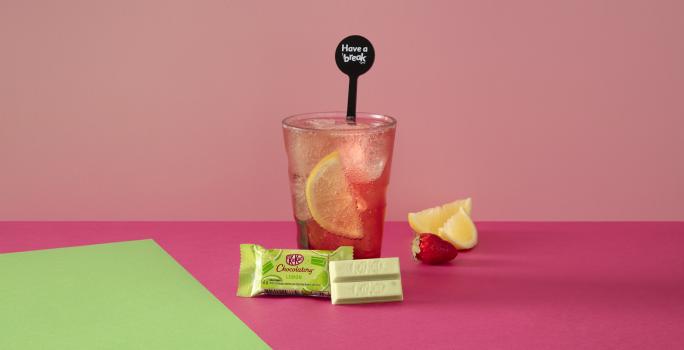 Foto da receita de Soda Italiana Pink Lemonade. Observa-se um fundo rosa e verde com um copo alto ao centro, servido cheio de gelo com uma rodela de limão siciliano dentro. À frente, um KIT KAT Limão.