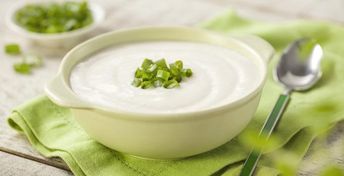 Fotografia em tons de verde em uma bancada de madeira com um guardanapo de pano verde ao centro com uma tigelinha branca e o creme de couve-flor dentro enfeitado com cebolinhas verdes. Ao lado, uma colher e ao fundo um potinho com cebolinhas verdes.