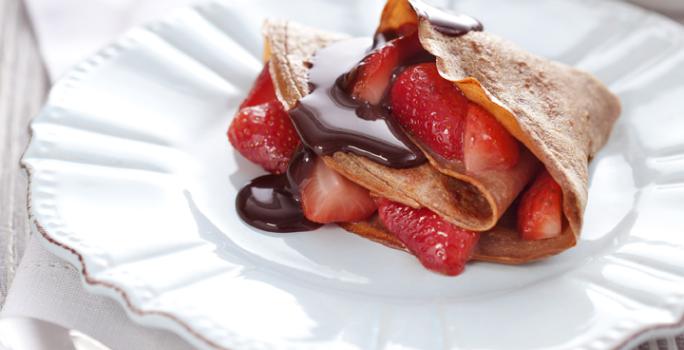 fotografia em tons de branco, marrom e vermelho de uma bancada cinza vista de frente, contém um prato branco redondo com um Crepe de Chocolate com Morangos e ao lado uma colher para servir.