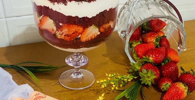 Foto em tons de vermelho da receita de red velvet na taça servida em uma graned porção em uma taça de vidro alta em cima de uma mesa de madeira com um pote de vidro com morangos ao lado