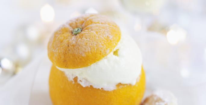 Fotografia em tons de branco com uma laranja inteira recheada de creme apoiada sobre um prato, em cima de uma mesa branca. Ao fundo, uma taça com champanhe.