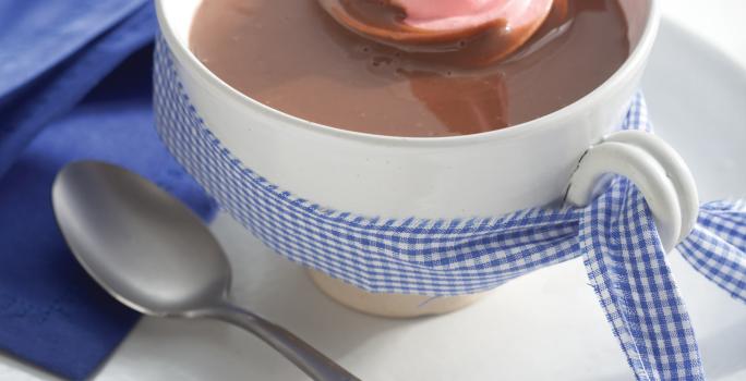Fotografia em tons de azul em uma bancada de madeira branca com um pano azul ao lado, um prato branco raso pequeno, um xícara branca e o chocolate quente com marshmallow dentro.