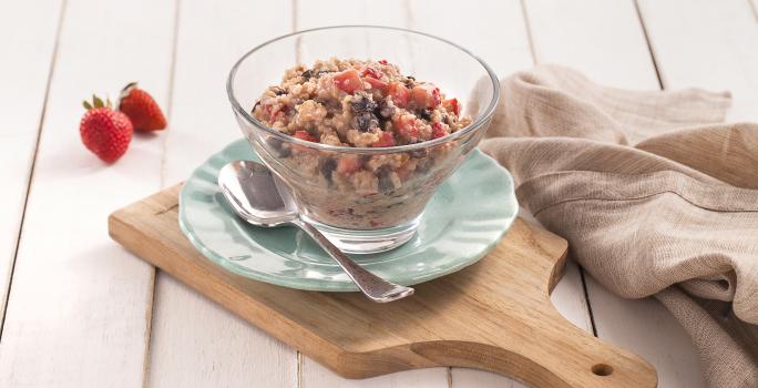 Fotografia em tons de azul em uma bancada de madeira clara, uma tábua de madeira, um paninho bege, um prato azul redondo pequeno, uma taça de vidro com o doce de quinoa com frutas secas, morango e iogurte dentro dele.