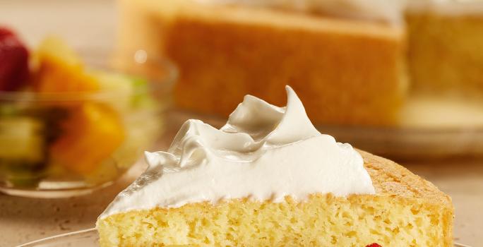 Fotografia em tons de vermelho em uma bancada com uma toalha branca com detalhes em flores vermelhas e folhas verdes. Ao centro, um prato de vidro com o pastel de tres leches com frutas.