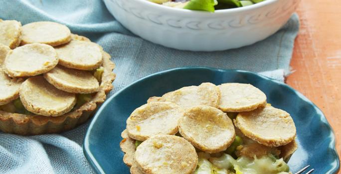 Fotografia em tons de azul em uma mesa de madeira laranja com um pano azul e um prato redondo fundo branco com salada e um prato fundo azul com a torta de massa de grão-de-bico com vegetais