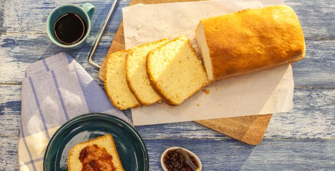 Fotografia em tons de azul e branco de uma bancada azul com uma tábua, sobre ela um paninho branco com o pão. Ao lado um paninho branco e azul, sobre ele um prato redondo azul com uma fatia de pão e geleia e uma xícara de café.