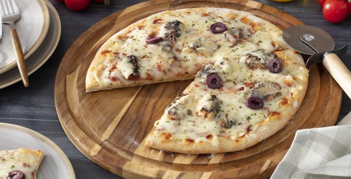 Foto da receita de pizza de sardinha servida em uma tábua de madeira circular com um dos pedaços separados em um prato de porcelana branco à frente. Para a decoração, há tomatinhos cereja, um paninho cinza, um vidro de azeite e um cortador de pizza