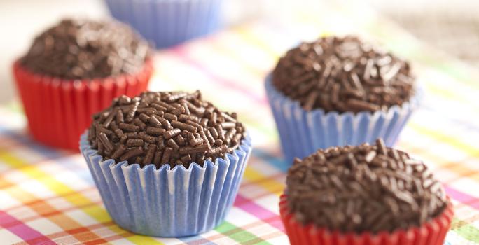 Fotografia em tons de azul e vermelho de uma grade com guardanapo quadriculado branco, verde, rosa, amarelo e azul com cinco brigadeiros e forminhas vermelhas e outras azuis. Ao fundo dois copos, um laranja e um azul.