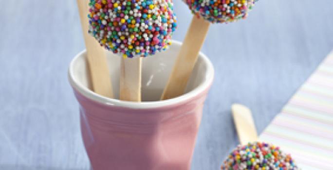 Fotografia em tons de lilás e azul, com copo lilás contendo três pirulitos de brigadeiro com confeitos coloridos, com mais um pirulito ao lado sobre guardanapo colorido, tudo sobre bancada em tons de azul.