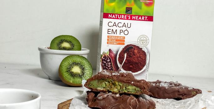 Foto em tons de verde e marrom da receita de kiwi com chocolate servida sobre uma mesa de mármore branco com kiwis e uma embalagem de cacau em pó nature's heart atrás.