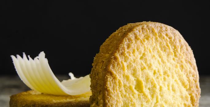 Fotografia de um biscoito em pé apoiado em outros dois biscoitos que estão atrás, e em cima desses que estão empilhados, tem uma lasca pequena de queijo. Estão sobre uma mesa de madeira em tons de cinza.