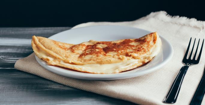 Foto aproximada da receita de Crepioca de Aveia em Flocos, em um prato branco, sobre um tecido em tom bege, com dois talheres de metal ao lado.