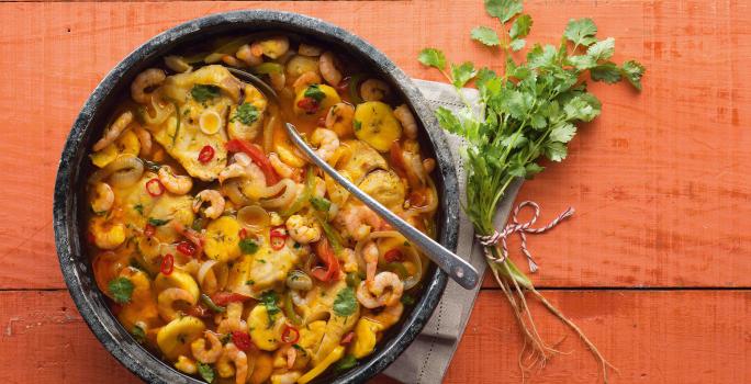 Fotografia em tons de vermelho em uma bancada de madeira vermelha com um recipiente redondo e fundo de pedra com a moqueca de peixe e camarão dentro dela.