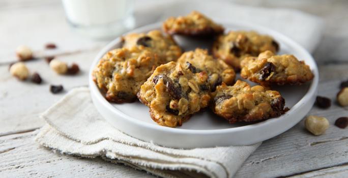 Fotografia em tons de branco, no centro, um prato branco com seis cookies com passas e grãos. Embaixo dos cookies, um pano de prato branco que está sobre uma mesa de madeira na cor branca.