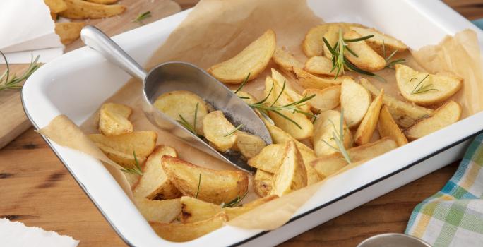 Fotografia em tons de amarelo e verde em uma bancada de madeira clara, um recipiente retangular com batatas feitas no forno e ramos de alecrim em cima dela. Ao lado, um pano xadrez em amarelo e verde.