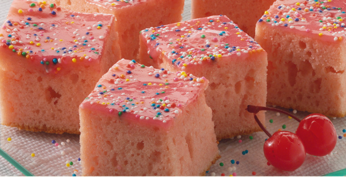 Vários pedaços de bolo rosa cortados em quadrados com confeitos coloridos por cima e duas cerejas ao lado.