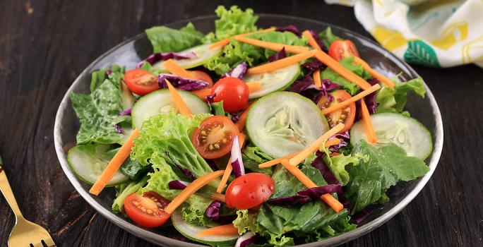 Fotografia de uma salada de folhas, pepino, cenoura ralada, tomates cereja e repolho roxo. Ao lado, um garfo na cor dourado, e ao fundo, uma tolha de mesa dobrada na cor branca, com desenhos em amarelo e verde, sobre uma mesa de madeira na cor preta.