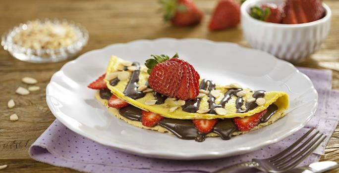 Fotografia em tons de lilás em uma bancada de madeira, com um paninho lilás com bolinhas, um prato branco raso com a crepioca recheada de chocolate e morangos.