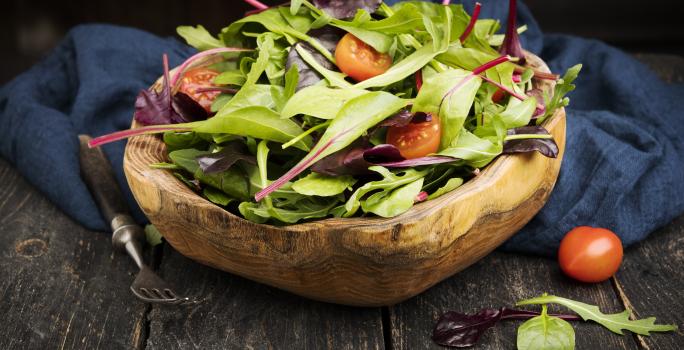 Fotografia em tons escuros de uma salada verde com salsão e tomates cereja dentro de um recipiente fundo de madeira, que está apoiado em um pano azul escuro, juntamente com um garfo de cabo preto, sobre uma mesa de madeira escura.