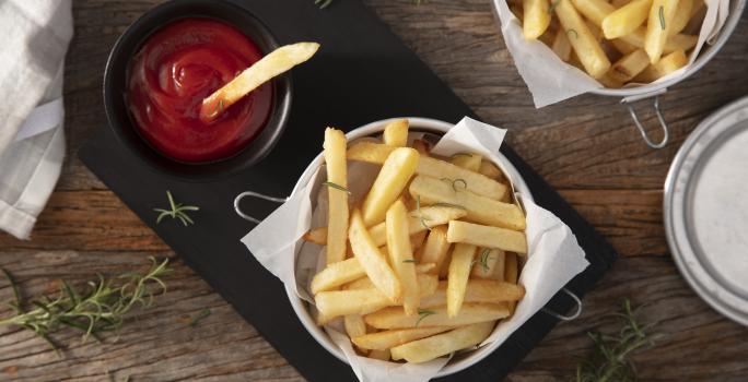 Fotografia em tons de marrom em uma bancada de madeira escura, uma tábua de ferro preta com um pote com batatas fritas dentro e um potinho ao lado com ketchup. Ao lado, um paninho e mais batata frita.