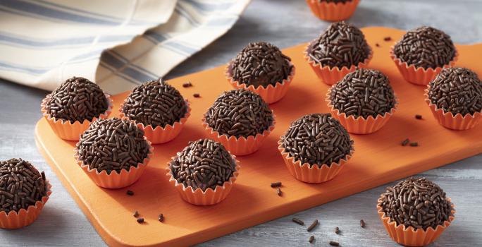 Imagem aproximada da receita de Brigadeiro Zero Lactose. Há diversos brigadeiros enrolados em forminhas em tom de laranja, todos sobre uma tábua de madeira em uma bancada decorada com um tecido em tom azul e bege