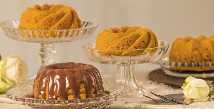 fotografia em tons de cinza, laranja e marrom vista de frente. Contém 4 bolos de cenoura e um deles esta com calda de chocolate, todos estão dentro de recipientes de vidros