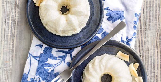 Fotografia em tons de azul em uma bancada de madeira clara, um pano branco com folhas azuis e dois pratinhos azuis escuros com o mini pudim de tapioca feito com iogurte, abacaxi e coco.
