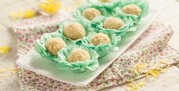 Fotografia em tons de verde em uma bancada de madeira com um recipiente branco retangular com os brigadeiros de torta de limão em forminhas verdes colocados em cima do recipiente. Ao lado, raspas de limão-siciliano espalhadas pela mesa.