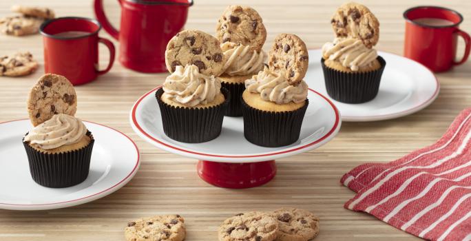 Foto de uma bancada clara com canecas e um bule vermelhos, um tecido listrado vermelho e branco. Há dois pratos brancos com cupcakes em cima e, ao centro, um prato com três cupcakes decorados com cookies.