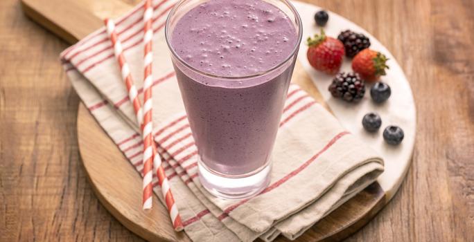 Foto em tons de roxo da receita de milkshake de uva com frutas vermelhas servida em um copo alto sobre uma tábua de madeira um pano listrado vermelho e branco com canudos de mesma cor e frutas vermelhas usadas na receita ao lado como decoração.