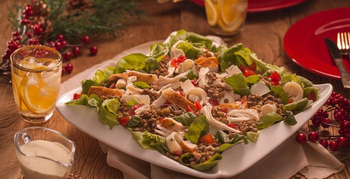 Fotografia em tons de vermelho em uma bancada de madeira escura, um pano bege, um prato branco grande com salada de alface, rúcula, lentilha e chester. Ao lado, copos, molho e enfeites e decoração natalina.