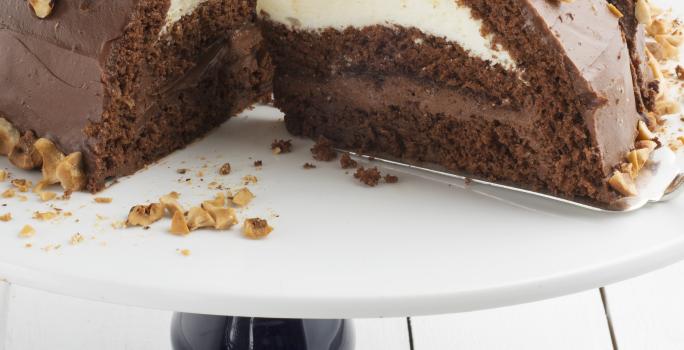 Fotografia em tons de marrom em uma bancada de madeira clara, um suporte de bolo branco com pé preto e uma doce de chocolate com recheio de creme branco e de chocolate.