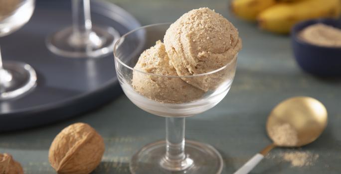 Foto da receita de Sorvete Crocante de Farinha Láctea Nestlé. Observa-se uma taça de sobremesa de vidro transparente com duas bolas do sorvete. A foto é decorada com Farinha Láctea, nozes com casca e bananas