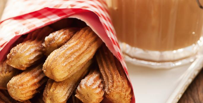 Fotografia em tons de marrom e branco de uma bancada de madeira com um prato retangular, sobre ele um paninho vermelho com churros. Ao lado um recipiente com doce de leite.