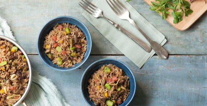 Fotografia em tons de azul em uma bancada de madeira azul com um pano branco com listras azuis, um panela com arroz e carne. Ao centro, dois potes redondos fundos com o arroz, carne e vegetais dentro dele.