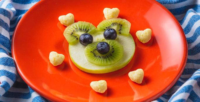 Fotografia em tons de laranja em uma bancada com paninho com listras azuis. Ao centro, um prato laranja redondo com um ursinho feito de maçã, kiwi e mirtilo e acompanhado por biscoitinhos em formato de coração. Ao lado, uma colher laranja e uma embalagem.