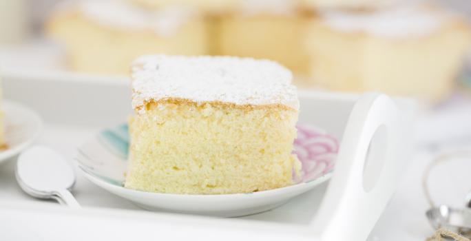 Fotografia em tons claros de um bolo de massa de pão de ló com açúcar de confeiteiro em cima, que está sobre um prato pequeno branco, com desenhos em rosa e azul. Ao fundo, mais pedaços do bolo, e ao lado do prato, colheres de café empilhadas.