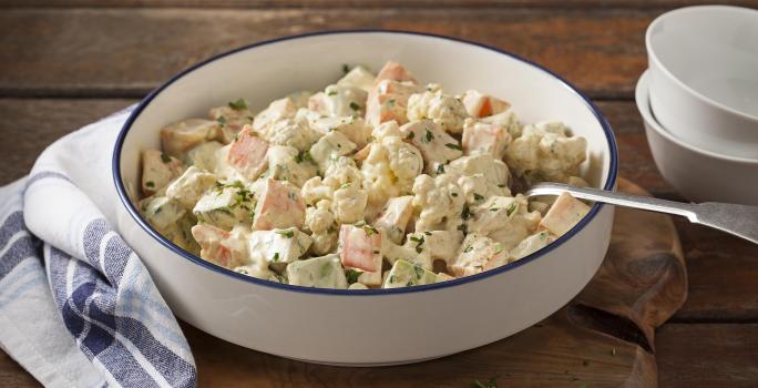 Fotografia em tons de marrom, branco e azul de uma bancada de madeira marrom com uma tábua de madeira, sobre ela um paninho branco xadrez com um recipiente redondo com a salada de legumes  e uma colher. Ao lado dois recipientes brancos.