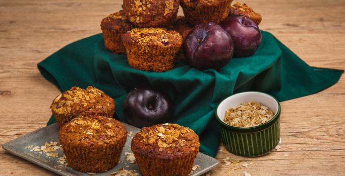 Fotografia em tons de verde em uma bancada de madeira clara, um pano verde, um recipiente retangular com os muffins de ameixa e cereais em cima. Ao lado, um potinho com cereais. Ao fundo, um suporte com o pano verde e mais muffins e ameixas.