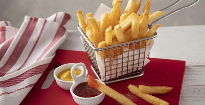 Fotografia em tons de vermelho de uma bancada branca, ao centro uma tábua vermelha com uma cestinha com batatas fritas dentro. Na frente um potinho com ketchup e um com mostarda.