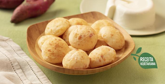 Fotografia em tons de verde e branco de uma bancada de verde clara com um recipiente de madeira clara com pães de queijo. Ao fundo um prato branco com o queijo minas e duas batatas-doces. Na frente um paninho branco.